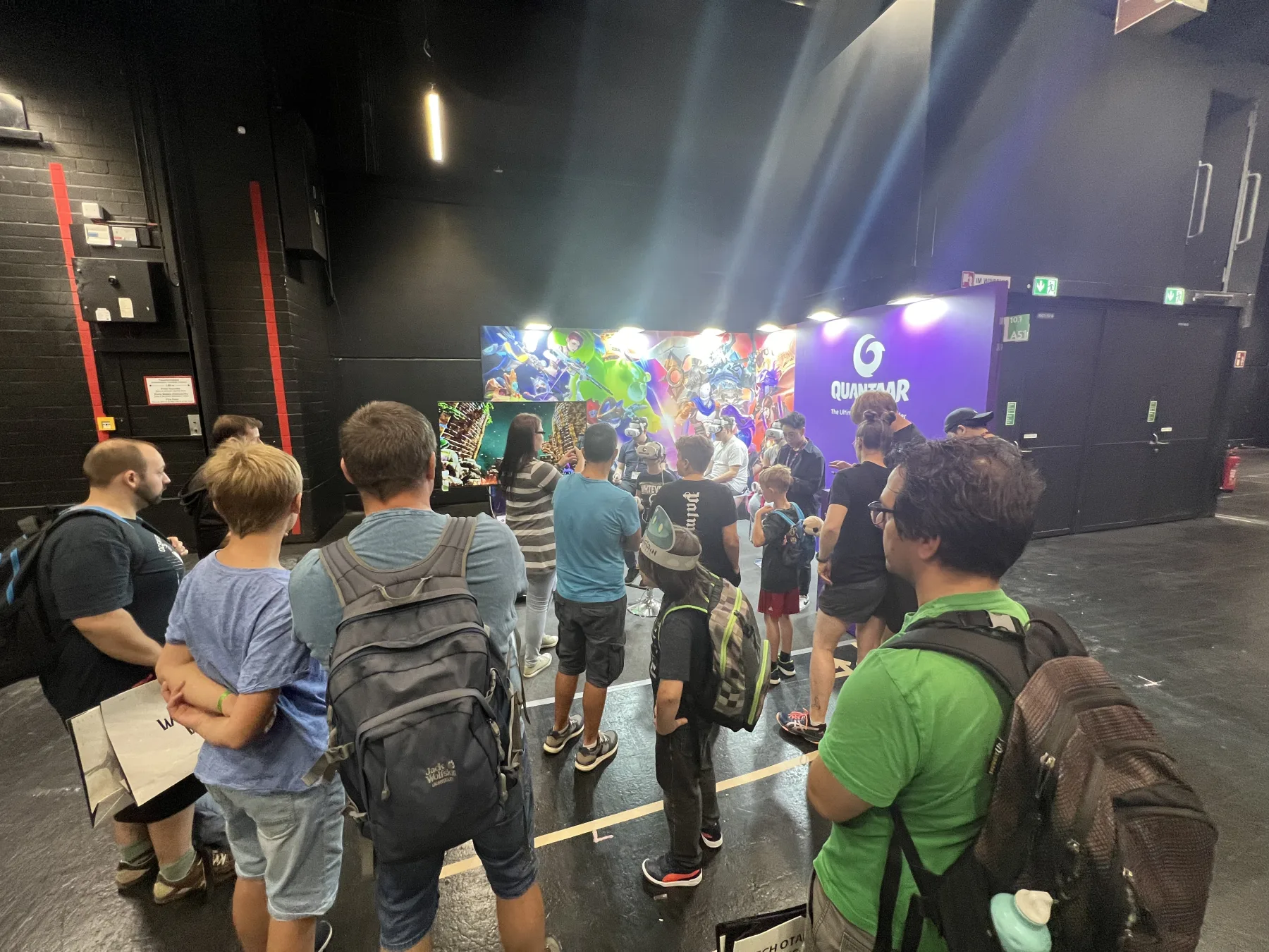 A dozen visitors with backpacks queueing in front of the Pumpkin VR QUANTAAR booth on the gamescom 2023 show floor in Cologne, the stand picked out by purple lighting and its character key-art wall