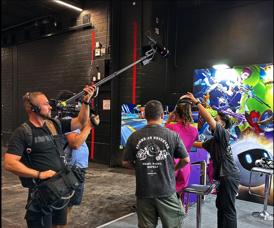 A camera operator with a shoulder-mounted camera and boom microphone filming at the Pumpkin VR booth at gamescom 2023 in Cologne, while staff help a visitor put on a VR headset beside the QUANTAAR key-art wall